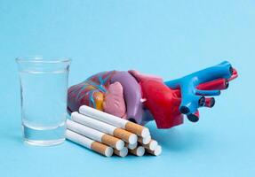 A glass of alcohol and a bunch of cigarettes with a heart organ on a blue background. The concept of bad habits badly affecting health and cardiovascular systems, close-up photo