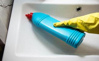The hands of a girl in a yellow cap with a blue bottle of detergent clean and wash the sink from limescale and dirt. Cleaning company services, close-up photo