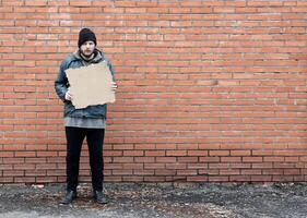 Homeless holds piece of cardboard. Refugee is at brick wall looking for job. Male tramp in dirty clothes. Below poverty line. Bearded fugitive beggar. Drunk immigrant. Copy space for text photo