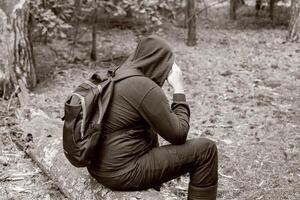A man with a backpack in a hood sits on a fallen tree in the middle of a clearing in the forest. Survival in the wild. Dehydration of the body. Mushroom picker lost in the forest. Black and white photo