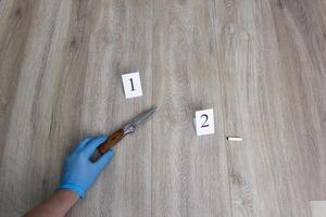 Criminalist in blue glove raises a collapsible knife from the floor, a cigarette butt on a wooden background, a close-up, an investigation photo