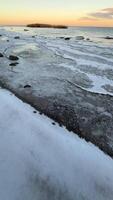 A serene winter sunset over a frozen lakeshore with scattered stones and bare trees in the background. The soft colors of the sky reflect on the icy surface, creating a tranquil and picturesque video
