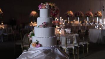Elegant Wedding Cake with Floral Decorations. A four-tier white wedding cake adorned with pink and peach flowers on a draped table. Warm lighting create a romantic reception atmosphere. video