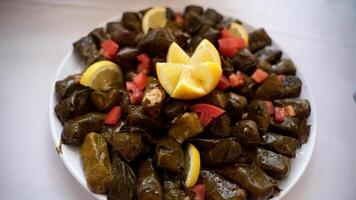 Plate of Stuffed Grape Leaves. Close-up of a white plate filled with stuffed grape leaves garnished with lemon wedges and diced tomatoes on a table. Mediterranean or Middle Eastern food concepts. video