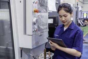 Asian woman industrial engineer wearing uniform looking and check of work on tablet with automated machinery in industrial workshop. photo