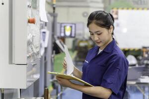 Asian woman industrial engineer wearing uniform checking of work list on clipboard with automated machinery in industrial workshop. photo