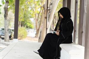 Side view of Muslim student wearing abaya niqab traditional clothes sit relaxing with tablet in university with sun fare and blurred background. photo