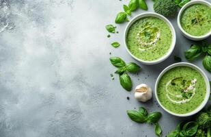 Fresh green basil soup in two bowls garnished with broccolini and garlic on a light grey surface surrounded by photo