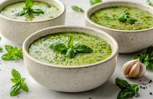 Fresh green basil soup in two bowls garnished with broccolini and garlic on a light grey surface surrounded by photo