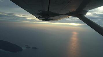 Airplane wing view from jetplane window on the sky in twilight sunset behind cloud at horizontal line and reflex golden orange light on sea wave surface. video