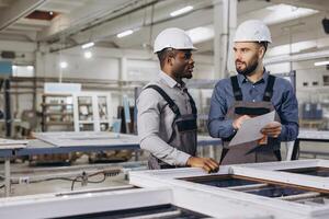 Engineers discussing blueprint while examining window frame in factory photo