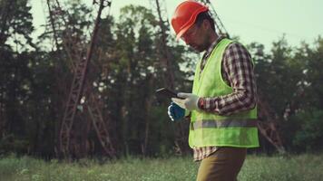 Engineer inspects high-voltage power lines, using tablet for calculations and planning. Dressed in safety helmet, he ensures integrity of electrical grid while monitoring transmission lines. video