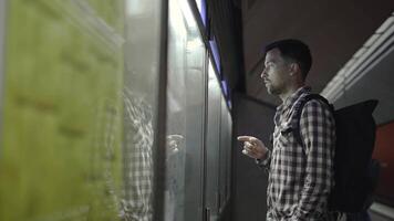 Man is looking at information on backlit board at public transport station in Germany, checking details on his smartphone. Passenger looks at subway train schedule at station on information board. video