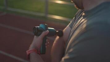 Athletic male massages muscles with hand massage gun, recovering from stadium running workout. . Body treatment with handheld wireless professional vibration shock massager after training session video