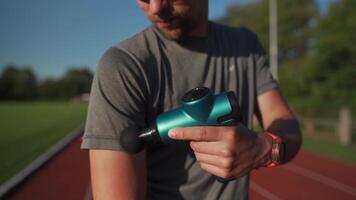 Athletic male massages muscles with hand massage gun, recovering from stadium running workout. . Body treatment with handheld wireless professional vibration shock massager after training session video
