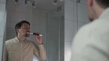 Caucasian man brushes his teeth with an electric toothbrush in the bathroom, view of the reflection through the mirror. Morning hygiene procedures. Oral irrigator. Morning routine for beautiful smile. video