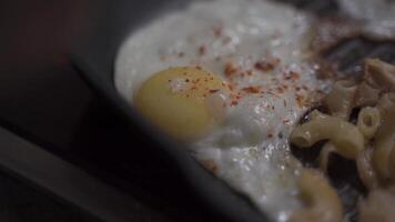 A close-up of hands seasoning a fried egg in a pan with noodles at home. The topic of cooking, unhealthy fried foods and cholesterol. Fried scrambled eggs and pan fried pasta with seasoning video