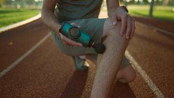 Athletic male massages muscles with hand massage gun, recovering from stadium running workout. . Body treatment with handheld wireless professional vibration shock massager after training session video