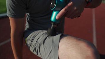 Athletic male massages muscles with hand massage gun, recovering from stadium running workout. . Body treatment with handheld wireless professional vibration shock massager after training session video