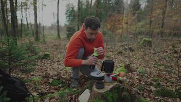 Man hiker pours water from flask into pot on gas burner on stump in woods to make tea and sublimated travel food. Boiling water on camping stove campfire in forest. Backpacking camping stove system. video