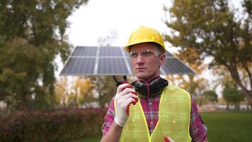 Male energy systems engineer walks on camera and talks on walkie-talkie in front of solar panel outside. Worker uses radio to communicate with colleagues in protective clothing at solar power plant. video