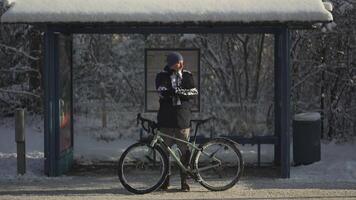 Cyclist extremely cold to ride bicycle in heavy frost and snow in winter and waiting for iced bus at stop, jumping and rubbing his hands. Snowy weather in Germany. A froze commuter on a bus stand. video