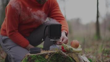 Man hiker boiling water on camping gas stove in woods on stump in fall. Camping utensils and gas burner. Traveler preparing meals in small pot with portable fuel gas torch on autumn camping trip. video