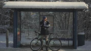 Frozen male cyclist waiting for bus at halt stop with bicycle in winter in snowy weather. Velocipede broke down and man waits long time for transport at the bus stop in winter after a snow storm. video
