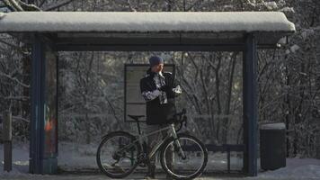 Frozen male cyclist waiting for bus at halt stop with bicycle in winter in snowy weather. Velocipede broke down and man waits long time for transport at the bus stop in winter after a snow storm. video