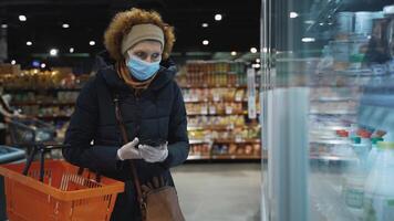 A woman shopper in a medical mask walks slowly along the shelves with the goods in search of the right ones, checking the list on the mobile phone.Safe shopping during a pandemic and an epidemic. video