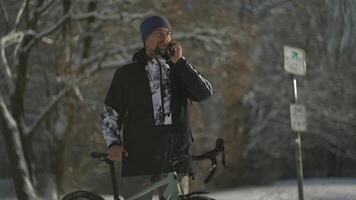 Male cyclist talking on mobile phone standing near bicycle on cycle path in snowy, sunny winter weather in Germany. Cycling in woods in winter. Active lifestyle and commuting on bicycle. ECO friendly. video