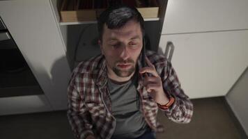 Drunken, depressed man sitting on the kitchen floor at home, drinking wine from a bottle and talking on his mobile phone. Sad male drinking alcohol while sitting on the floor and making phone calls. video
