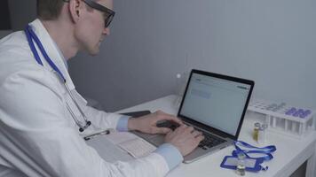 Young male physician in glasses and white lab coat working with paperwork sitting at desk operating laptop in a doctors office. Doctor reviews patients labs and input data into computer. video