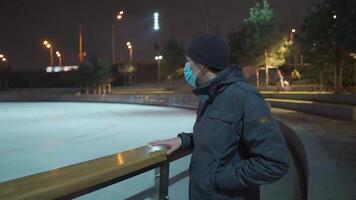 Masked male stands near skating rink arena with decorated Christmas tree empty during quarantine, coronavirus and covid pandemic in evening. Skating rink at night. An empty public ice skating area. video