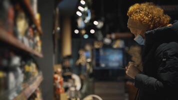 A woman shopper in a medical mask walks slowly along the shelves with the goods in search of the right ones, checking the list on the mobile phone.Safe shopping during a pandemic and an epidemic. video