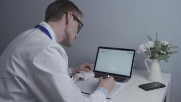 Male doctor working at computer in private medical practice, checking patients tests and making notes in notebook. General medicine doctor working with documents in clinic in glasses and white coat. video