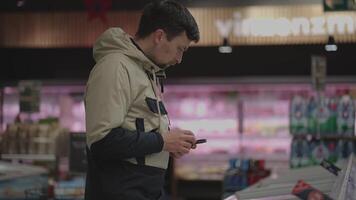 Man with smartphone choosing frozen food from supermarket freezer. Male reads product information on phone while standing at refrigerators in market. Shopping list on a smartphone. Frozen products. video