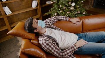 Young man in mask lies on sofa and stares at ceiling, suffering from loneliness and home isolation during Christmas holidays, coronavirus quarantine. Male suffering from boredom looking up covid 19. video