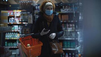 A woman shopper in a medical mask walks slowly along the shelves with the goods in search of the right ones, checking the list on the mobile phone.Safe shopping during a pandemic and an epidemic. video