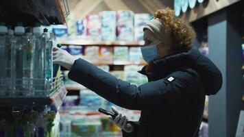 A housewife in supermarket selects products from a list, looks mobile phone and searches for product.The topic of safety in society during a pandemic, use of medical protection, a mask on the face. video