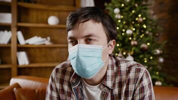 Young upset with bad mood man in protective mask is suffering from loneliness at home while sitting on couch near Christmas tree during New Year holidays. New norm, lockdown and quarantine coronavirus video
