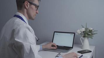 Male doctor working at computer in private medical practice, checking patients tests and making notes in notebook. General medicine doctor working with documents in clinic in glasses and white coat. video