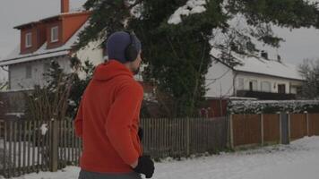 Male runner jogging in winter in snow covered park in Europe and listening to music on large headphones rear and side view. Man jogger running in snowy park and listen to music with big earphones. video