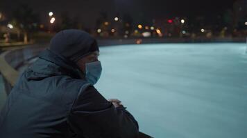 A sad man in a mask looks at an indoor ice arena with a Christmas tree in the center of the rink in the evening during lockdown, coronavirus pandemic. Empty public place for skating. video
