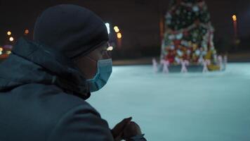 A sad man in a mask looks at an indoor ice arena with a Christmas tree in the center of the rink in the evening during lockdown, coronavirus pandemic. Empty public place for skating. video