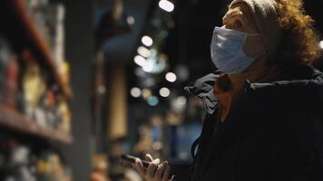 An older woman in grocery store chooses goods while examining in her hands, wearing a medical mask during the coronavirus epidemic, maintains a social distance. The topic is health safety in society. video