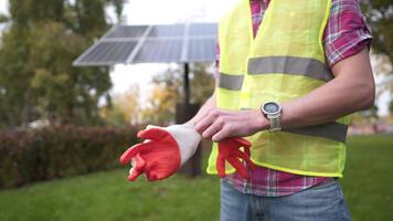 Engineer on workplace puts on protective gloves while standing in front of solar panel. Technician wearing work gloves in front of sun panels. Green clean energy. Worker putting on work gloves. video