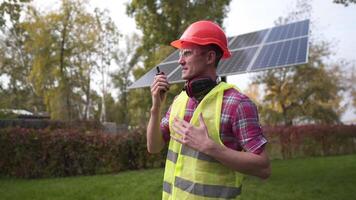 Caucasian male worker in a red helmet, green vest and protective equipment serves a small solar panel. The engineer talks and gives commands over the radio at the solar power station. video