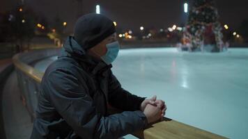 Masked male stands near skating rink arena with decorated Christmas tree empty during quarantine, coronavirus and covid pandemic in evening. Skating rink at night. An empty public ice skating area. video
