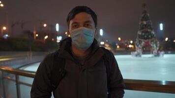 A sad man in a mask looks at an indoor ice arena with a Christmas tree in the center of the rink in the evening during lockdown, coronavirus pandemic. Empty public place for skating. video